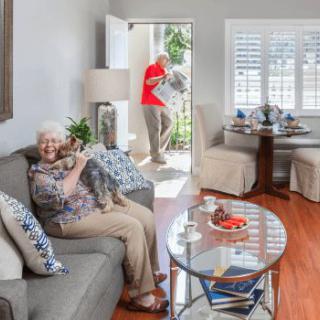 An elderly woman sitting on a sofa with a cat in her lap, as someone tends to the plants outside.