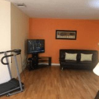 A cozy living room with an orange accent wall, featuring a treadmill, television, and black sofa.