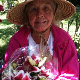A woman wearing a straw hat and pink jacket holds a bouquet of flowers in a garden.