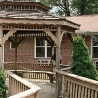 A wooden gazebo with a ramp leading to a brick building surrounded by greenery.