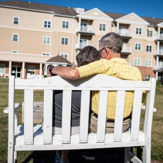 An elderly couple sits closely on a bench outside a retirement home.
