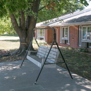 A swing bench under a large tree on a shaded patio in front of a house.