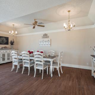A spacious dining room with a large table and chairs, adorned with chandeliers and a ceiling fan, featuring wood flooring and decorative elements.