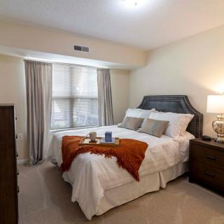 A cozy bedroom with a neatly made bed, featuring pillows and an accent blanket. Natural light filters through the curtains, and bedside lamps illuminate the space.