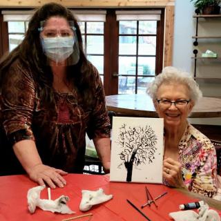 Two women enjoying a painting activity indoors, one holding a completed artwork of a tree.