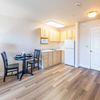 A compact kitchen and dining area featuring wooden cabinetry, a small round table with chairs, and bright natural lighting.