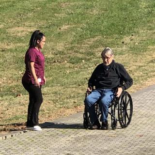 A caretaker interacts with an elderly person in a wheelchair on a sunny day.