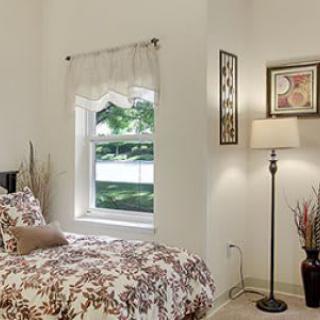 A cozy bedroom with a floral bedspread, standing lamp, wall art, and natural light streaming through the window.