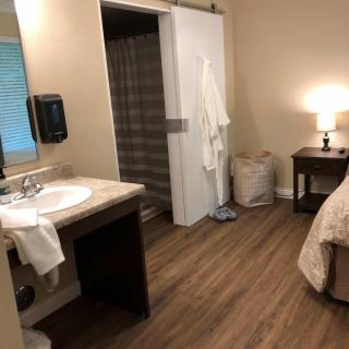 A cozy bedroom with an adjoining bathroom, featuring a bed, bedside table with a lamp, and a sink area.