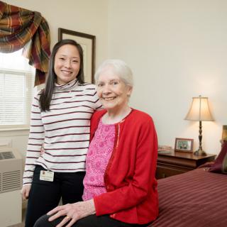 A young woman stands beside an elderly woman seated on a bed, both smiling warmly in a cozy bedroom setting.