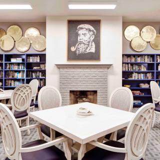A cozy library with bookshelves, a central table with chairs, and a fireplace adorned with a portrait.