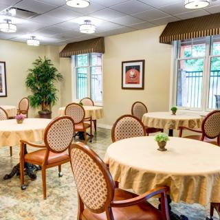 A cozy dining room with round tables, chairs, and indoor plants, well-lit with natural light from the windows.