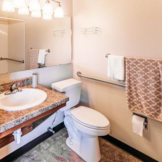 A clean and well-lit bathroom with granite countertops, featuring a sink, toilet, mirror, towel rack, and accessibility features.
