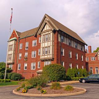 A large brick building with a distinctive roofline and a flagpole in front, surrounded by landscaped greenery.