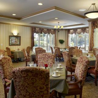 A cozy dining room with elegantly set tables and upholstered chairs, featuring warm lighting and classic decor.