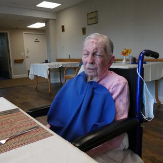 An elderly person sitting in a wheelchair at a dining table.
