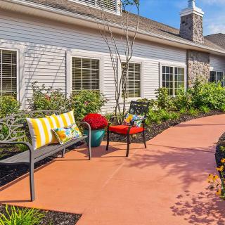 A cozy outdoor patio area with benches and chairs, adorned with colorful pillows, surrounded by a landscaped garden.