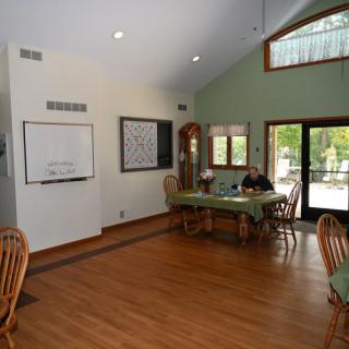 A spacious dining area with wooden flooring, a central table and chairs, illuminated by natural light from large windows.