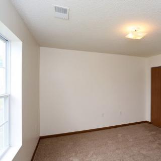 An empty room with carpeted flooring and a large window, featuring a ceiling light fixture and a wooden door.
