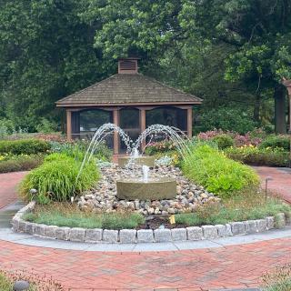 A beautiful garden with a central fountain surrounded by lush greenery, and a gazebo in the background.