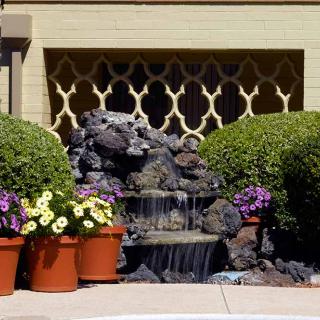A small garden with a cascading fountain surrounded by potted flowers and trimmed bushes.