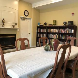 A cozy dining room with a large table, surrounded by wooden chairs, and a bookshelf along the wall. A vase sits in the center of the table, and a fireplace adds warmth to the setting.
