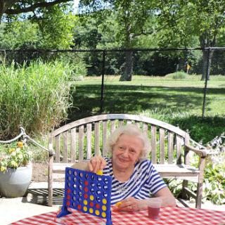 An elderly woman enjoys a game of Connect Four on a sunny day, surrounded by nature.