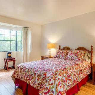 A cozy bedroom featuring a floral patterned bedspread, natural light from a large window, and warm wooden furniture.