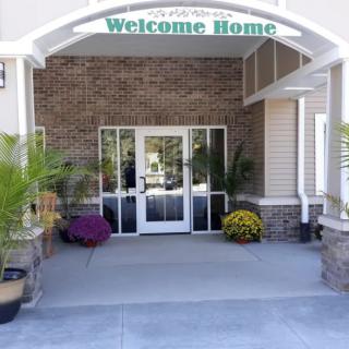 A welcoming building entrance with potted plants and a 'Welcome Home' sign above the door.