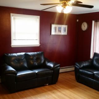 A cozy living room with maroon walls, featuring black leather couches, a ceiling fan, and wooden flooring.
