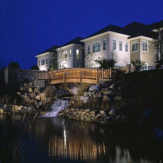 A beautifully lit house at night with a wooden bridge over a small waterfall.