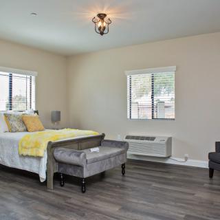 A spacious bedroom with hardwood flooring, featuring a bed, seating area, and natural light from two windows.