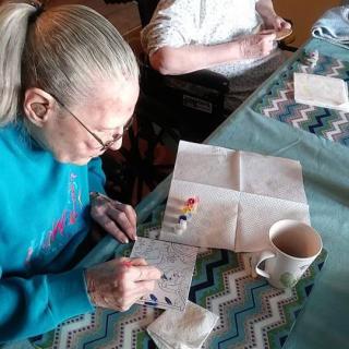 An elderly woman engaged in painting at a table, with a mug nearby.
