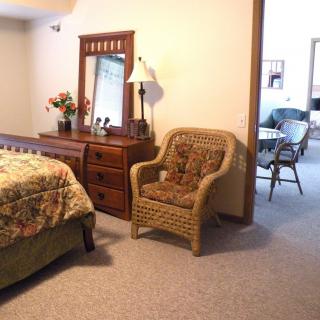 A cozy bedroom with a wicker chair, wooden dresser with mirror, and a bed with patterned bedding.