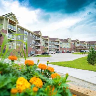 A vibrant apartment complex with colorful flowers and a spacious parking area under a cloudy sky.