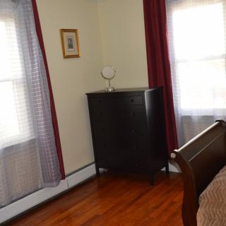 A cozy bedroom corner featuring a dark dresser, a small lamp, and two curtained windows letting in natural light.