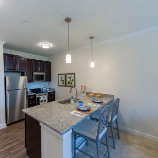 A modern kitchen with a granite countertop and stainless steel appliances, featuring pendant lighting and bar stools.