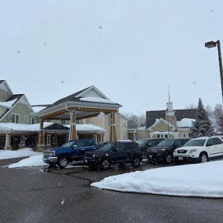 A snowy day at a community center with vehicles parked out front and a church in the background.