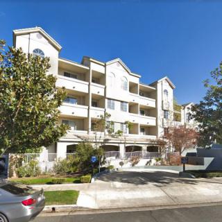 A modern apartment building with a driveway, surrounded by trees and parked cars on a sunny day.