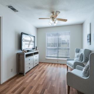 A modern living room with wooden flooring, featuring a television, chairs, and natural light from a large window.