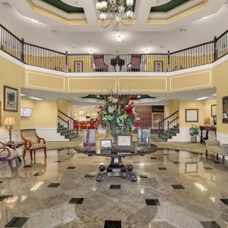A grand hotel lobby featuring a luxurious interior with a central floral arrangement, elegant staircase, and marble flooring.