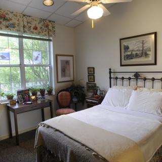 A cozy bedroom with a neatly made bed, artwork on the walls, and a window letting in natural light.