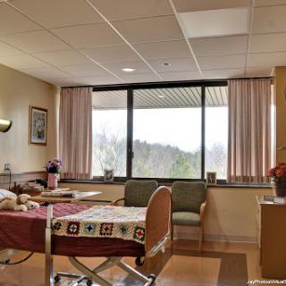 A brightly lit hospital patient room featuring a bed, seating area, and large window overlooking a scenic view.