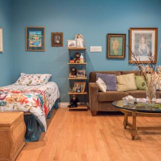 A cozy living space featuring a bed with a floral duvet, a comfortable sofa with cushions, and artwork adorning the blue walls.