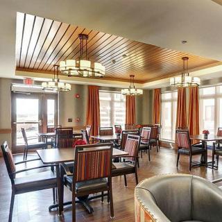 A well-lit dining room with wooden ceilings, featuring multiple tables and chairs, elegant chandeliers, and large windows with curtains.