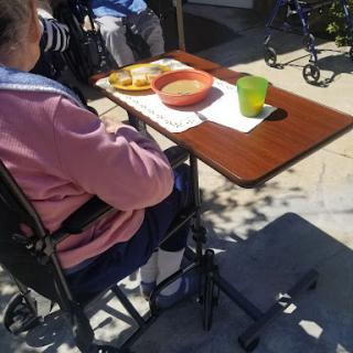 An elderly individual enjoying a meal outdoors, seated in a wheelchair with a tray of food and a drink.