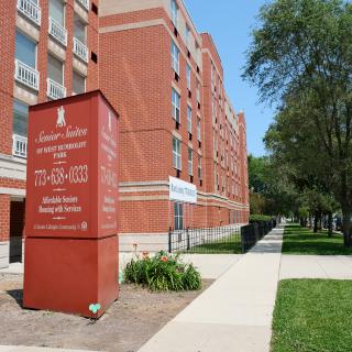 A red brick university building with a sign on the campus, surrounded by trees and a sidewalk.