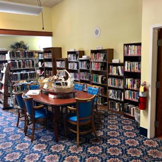 A cozy library corner with shelves filled with books surrounding a table and chairs on a patterned carpet.