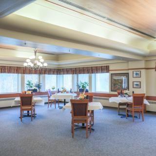 A spacious dining room with several tables and chairs, large windows, and plants decorating the space.