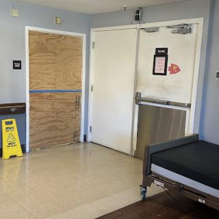 A hospital room with a closed door that appears to be under maintenance, featuring a bed and a caution sign on the floor.